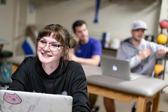 Students are smiling, seated at desks in a classroom with their laptops in front of them