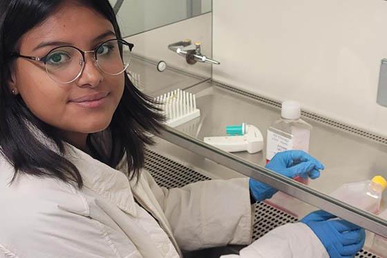 Photo of a young woman with glasses, wearing a lab coat and blue latex gloves, poses for a photo while working in a lab