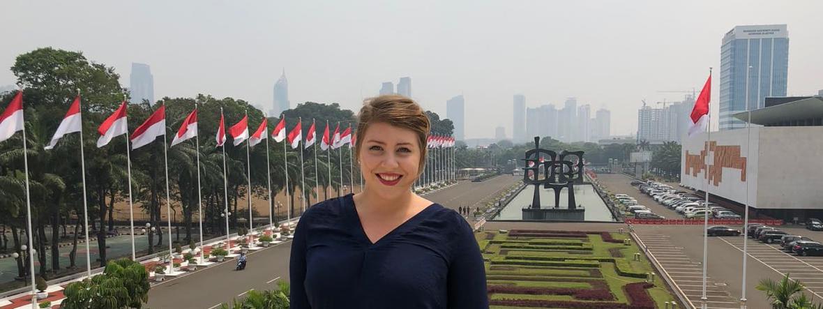 Photo of a female Chatham University student posing outdoors in front of a city skyline and a gallery of flags. 