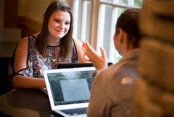 Photo showing the back of a young woman speaking to a peer, who is paying attention to the conversation. There is a computer and work on a table between them.